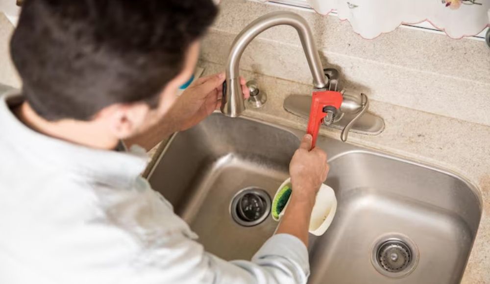 Muškarac pomoću crvenog vodoinstalaterskog ključa proverava slavinu iznad duple kuhinjske sudopere od nerđajućeg čelika